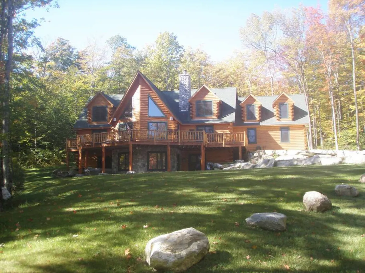 This image showcases the exterior of a classic Vermont log cabin nestled among mature trees, featuring a spacious deck and a prominent stone chimney. The surrounding greenery highlights the natural setting, while the open deck hints at opportunities for outdoor gatherings and relaxation.