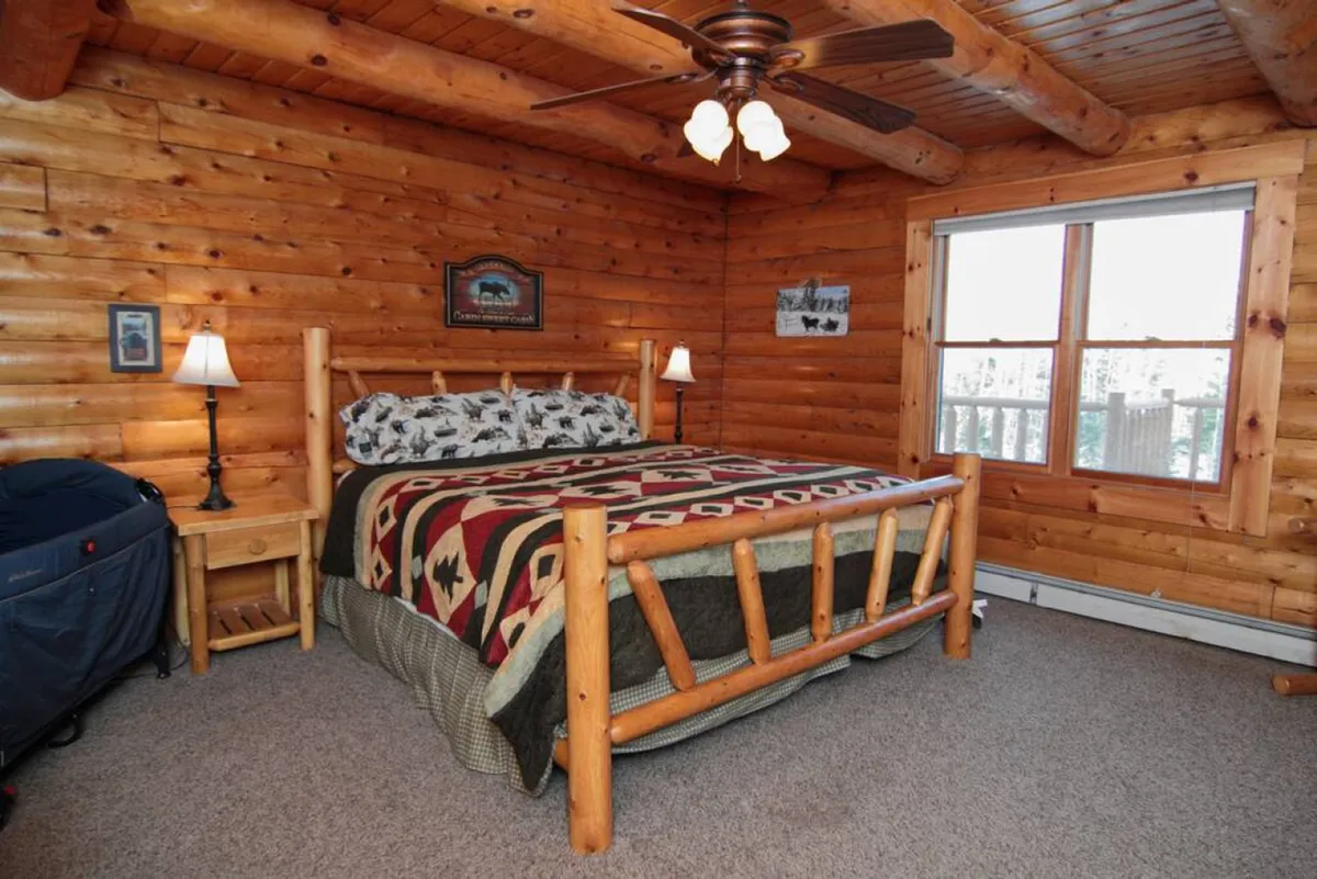 A serene bedroom with a rustic log bed positioned beneath a large window, allowing sunlight to filter in. A decorative blanket and matching nightstands create a cozy, restful atmosphere perfect for unwinding. The view outside hints at the wooded surroundings.
