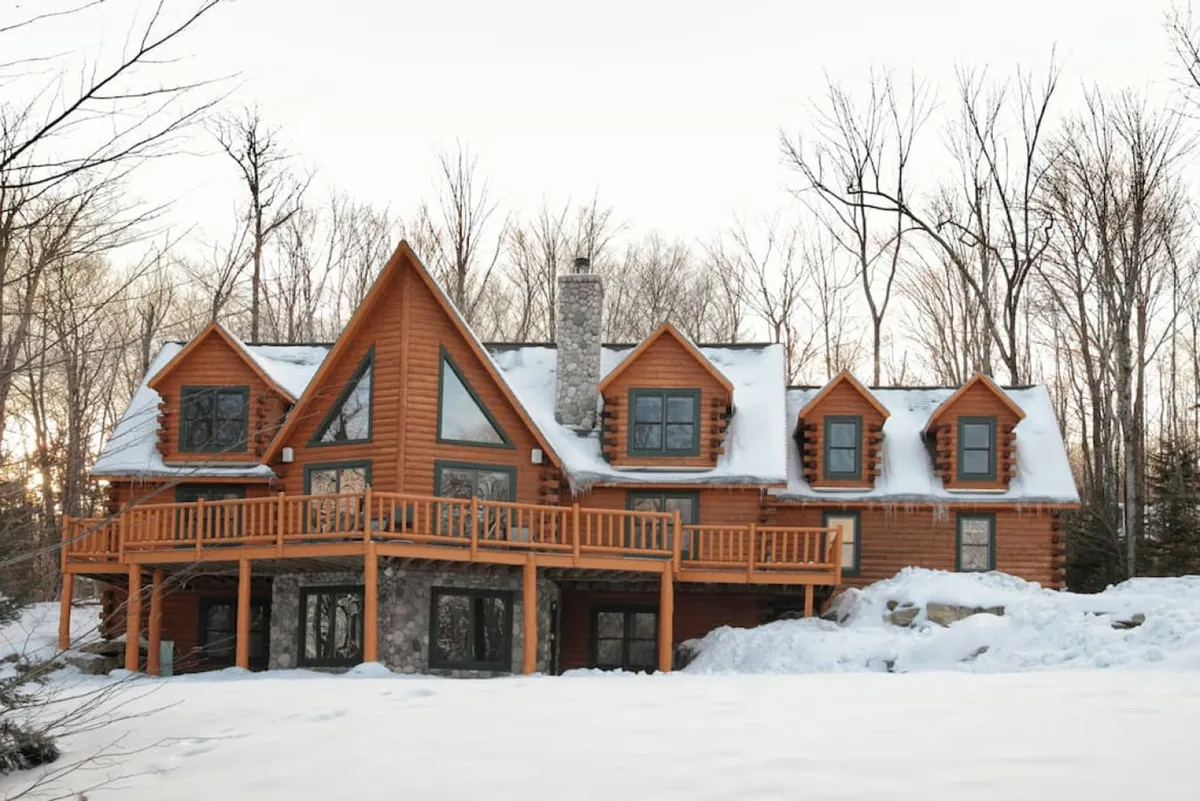 The image showcases a grand hand-peeled cedar log cabin with a welcoming stone chimney, expansive deck, and charming dormer windows, set amidst a beautifully landscaped Vermont yard. The scene evokes warmth and comfort, hinting at cozy gatherings, relaxation, and outdoor enjoyment in any season.