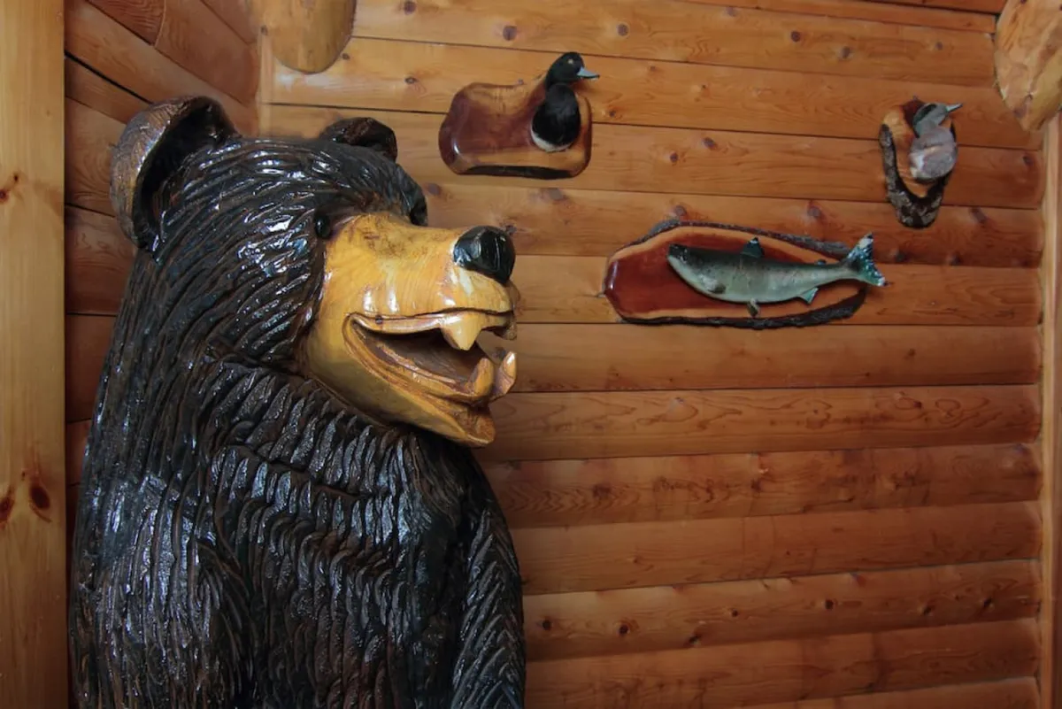 A charming corner showcasing a wooden bear carving along with fish and bird carvings set against a log wall. The cozy decor creates a nature-inspired ambiance, ideal for group photos and storytelling sessions during festive gatherings.