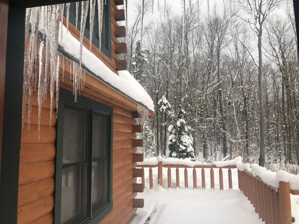 This image features the log cabin's exterior enveloped in fresh snow, with icicles hanging from the roof and a snow-covered deck that hints at recent snowfall. The scene is tranquil, encapsulating the quintessential winter retreat atmosphere, and visually connects the property to the surrounding winter landscape.