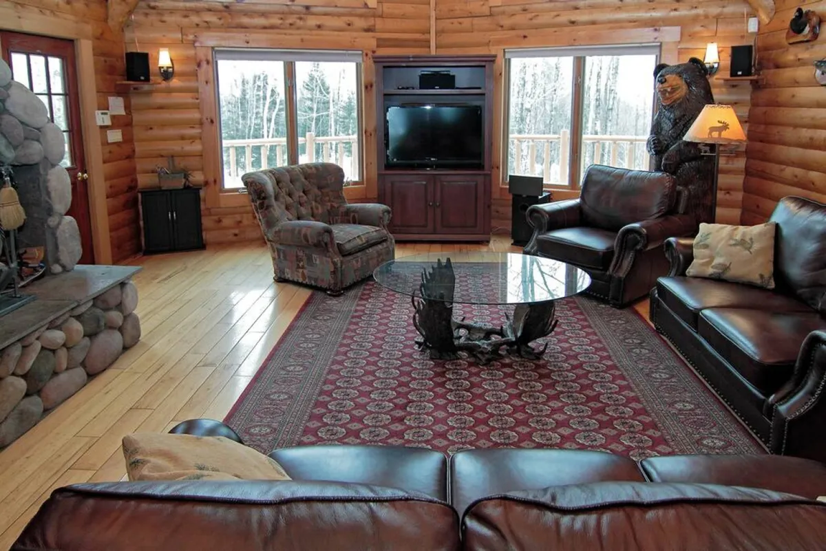 Featuring comfortable seating clustered around a stone fireplace, floor-to-ceiling windows revealing the outdoors, and a spacious coffee table, this image radiates warmth and openness. Family members can be imagined relaxing, sharing stories, or enjoying the fire after outdoor adventures.