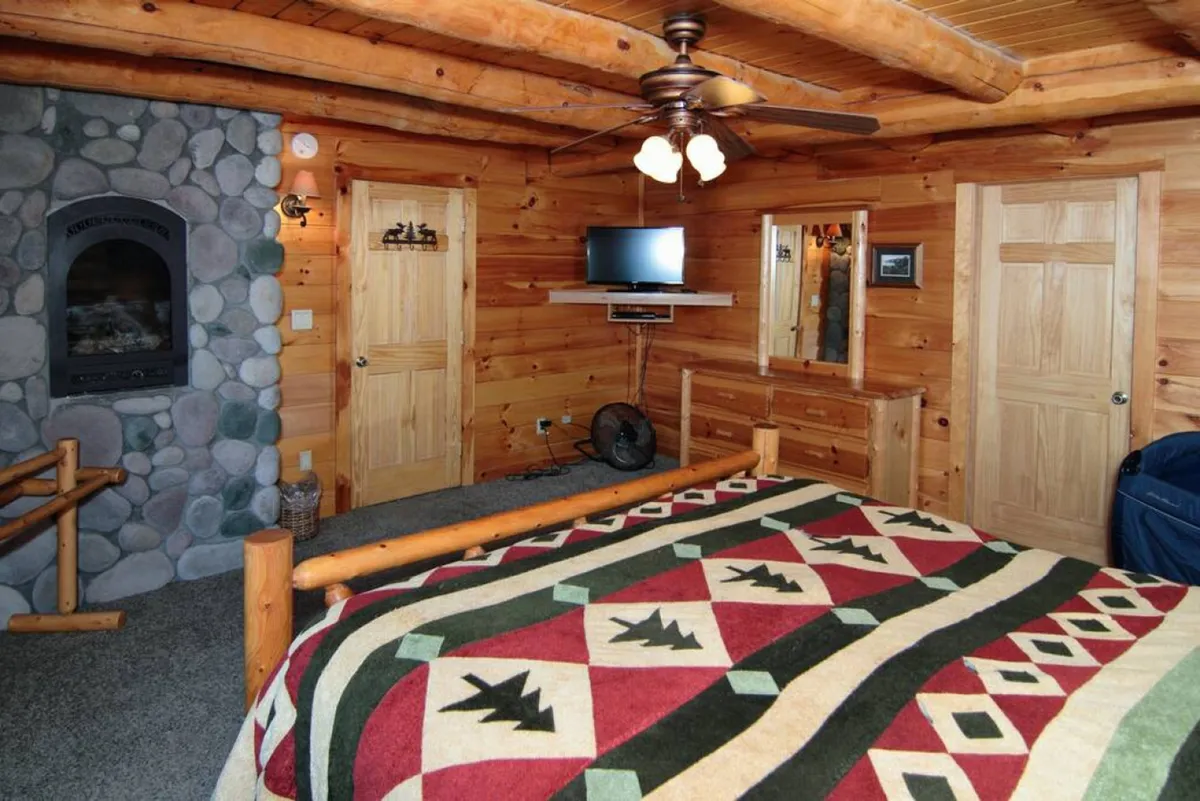 This inviting room features a cozy bed adorned with a patterned blanket, a striking stone fireplace, exposed wooden ceiling beams, and a television mounted on the wall. The space radiates warmth and comfort, perfect for gathering groups to relax, watch movies, or share stories around the fire.