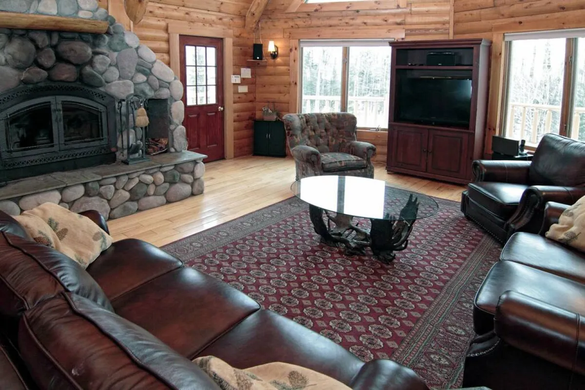 This image showcases the Great Room with a dramatic stone fireplace rising to cathedral ceilings, exposed wooden beams, large windows flooding the space with natural light, and plush leather sofas arranged around a decorative rug. The scene is set for family gatherings, cozy reading sessions, and lively game nights, emphasizing comfort and rustic elegance.