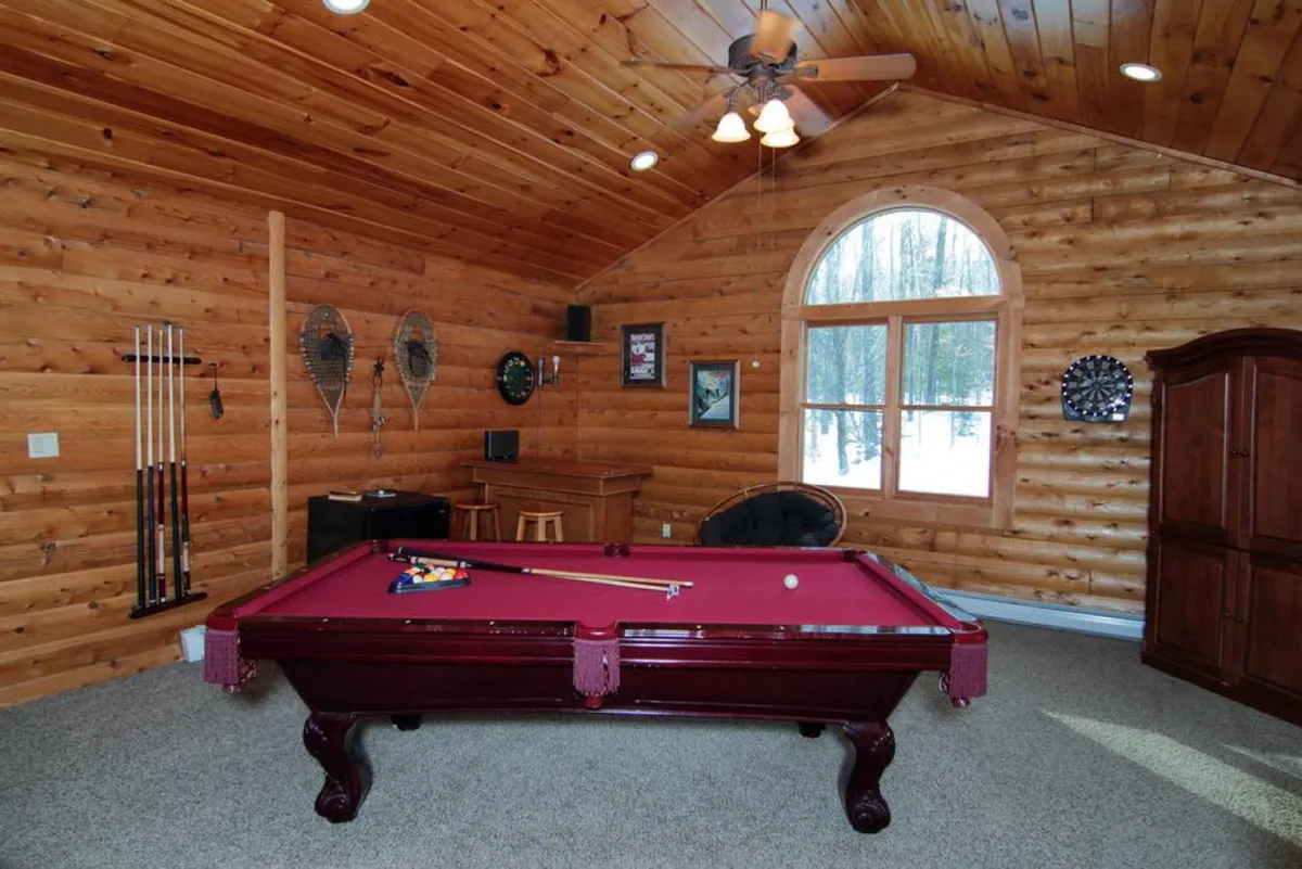 A spacious loft area featuring a pool table as the central focus, surrounded by wooden walls and large windows that allow natural light to flood the room. Comfortable furniture is arranged nearby for relaxation and socialization, creating a warm and inviting atmosphere.