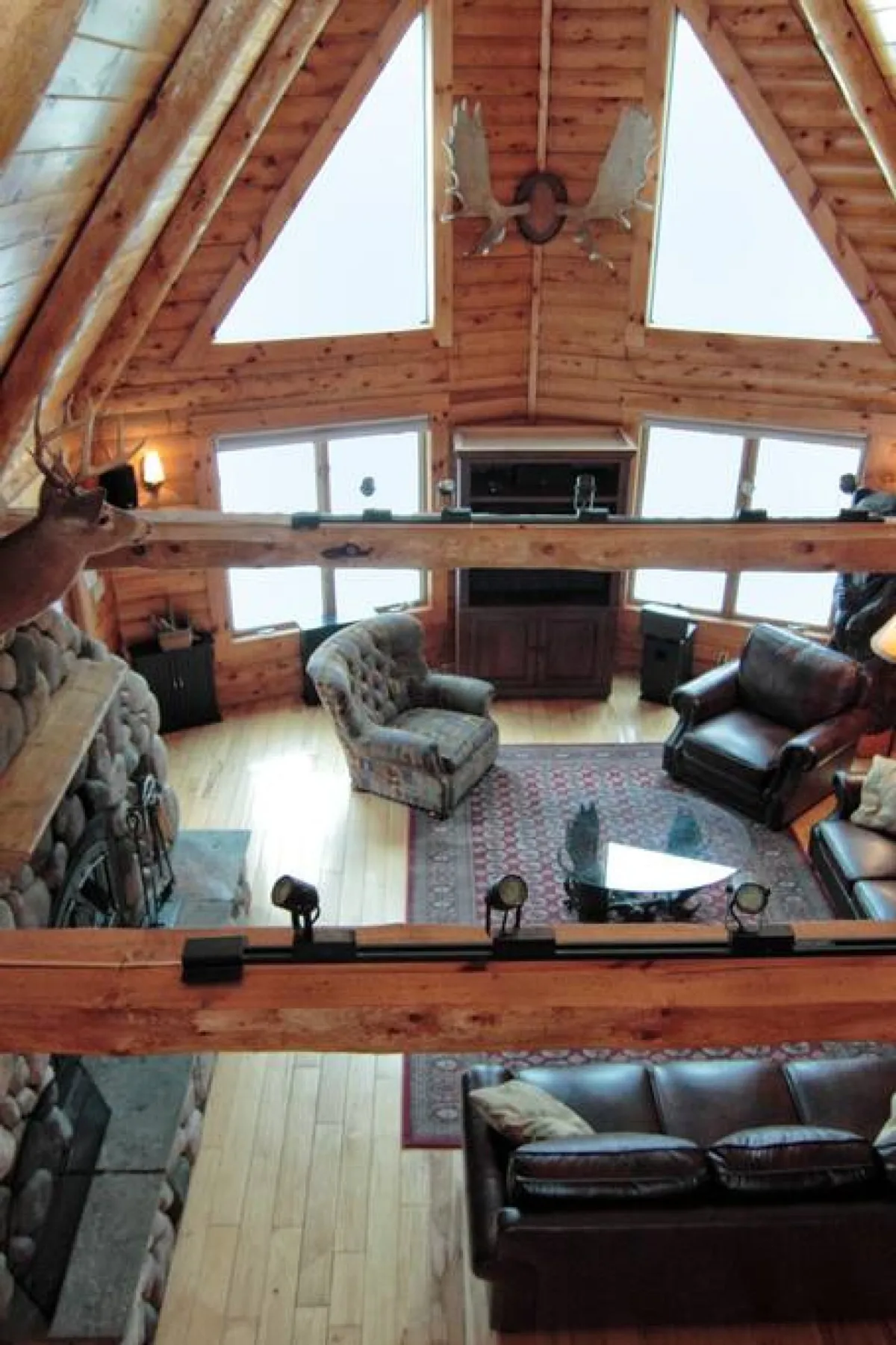 The Great Room is depicted from a different angle, highlighting the expansive stone fireplace, large windows drawing in sunlight, exposed wooden beams, decorative antlers, and ample comfortable seating. The atmosphere is warm and inviting, ideal for sharing stories, enjoying quiet reflection, or hosting lively game nights.
