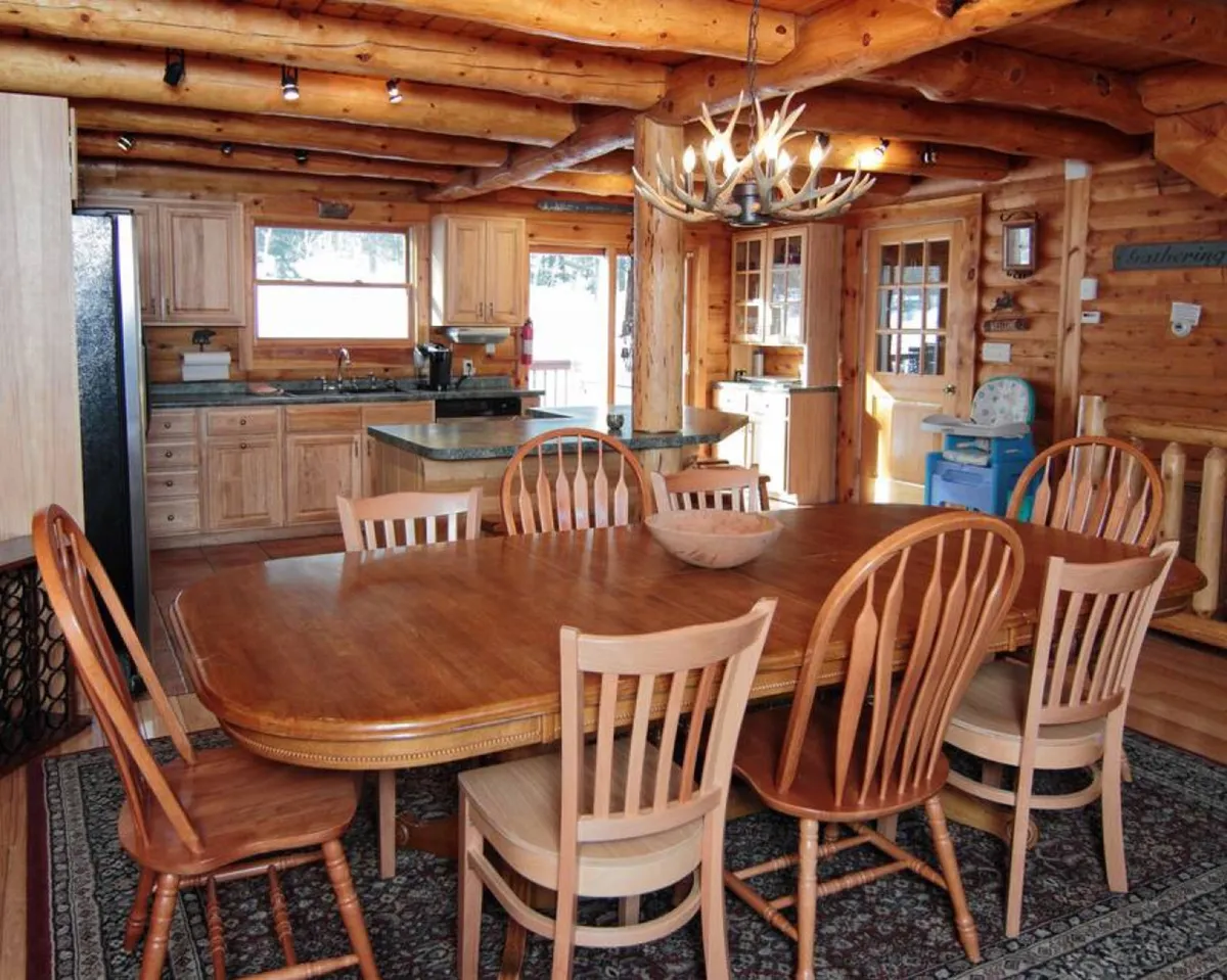 A spacious, warmly-lit dining area featuring a large wooden table surrounded by chairs, topped with a rustic antler chandelier. The kitchen area is visible in the background, suggesting an open-plan space ideal for both preparing and sharing meals.